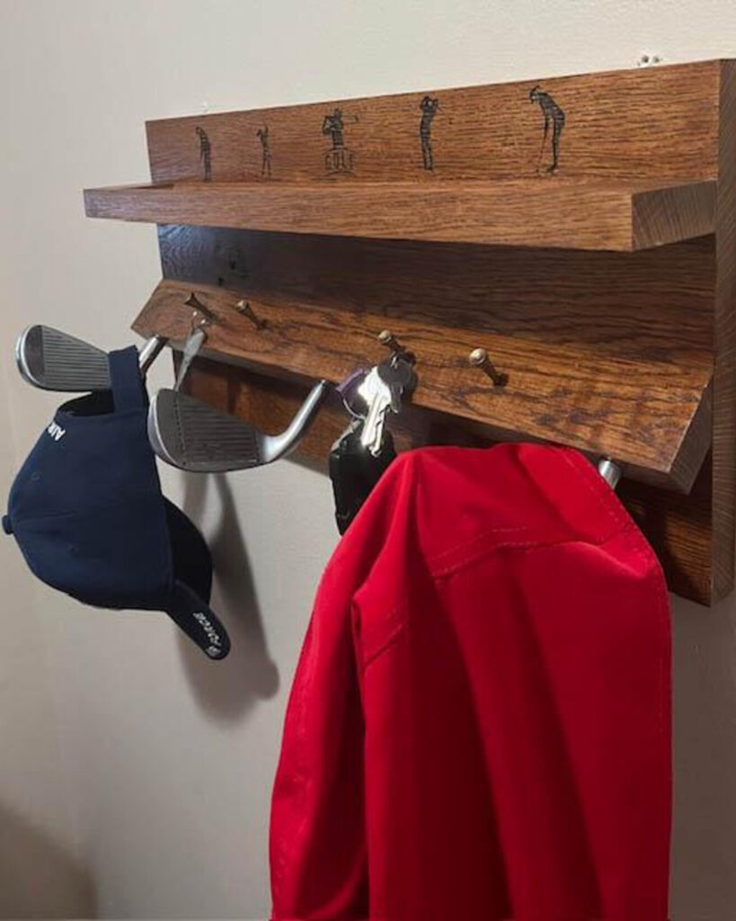 A wooden coat and hat rack featuring upcycled tees and golf clubs as hooks. There is extra shelf space above the hooks, with engraved images of golfers in various poses along the back panel.